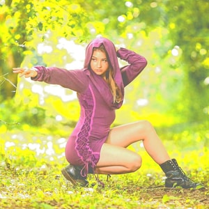 May include: A purple hooded dress with lace-up detailing and black boots. The image is set outdoors with a blurred green and yellow background.