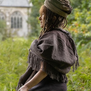 May include: A dark brown shawl with a fringed edge, worn over the shoulders. The person is wearing a turban-style head covering. The background features a green field and a building with arched windows.