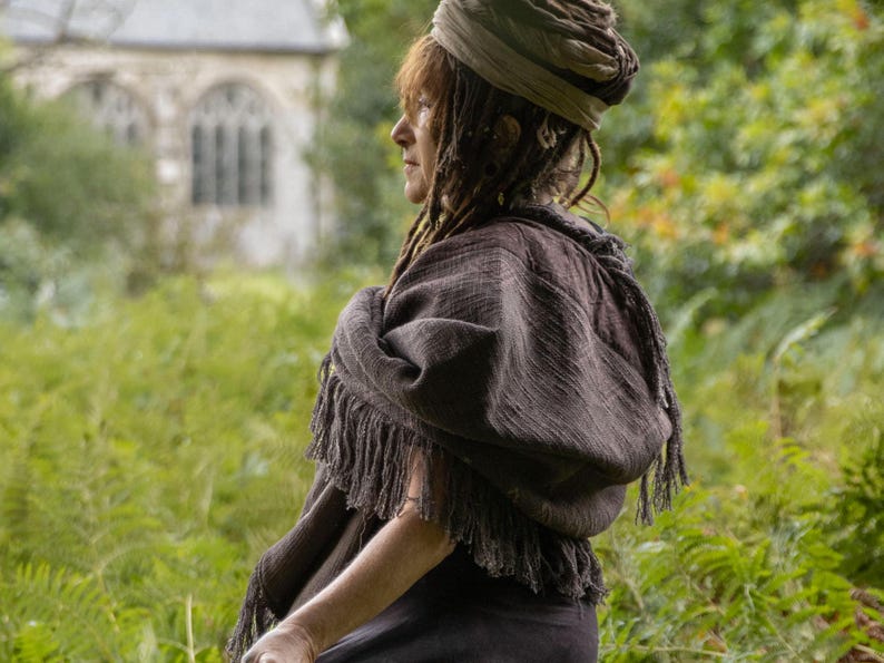 May include: A dark brown shawl with a fringed edge, worn over the shoulders. The person is wearing a turban-style head covering. The background features a green field and a building with arched windows.
