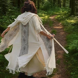 May include: Cream-colored hooded robe with intricate black geometric patterns along the front and sleeves. The robe has a fringed hem and is worn outdoors on a forest path. The person is holding a wooden staff.