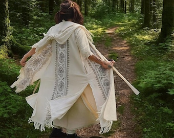 Könnte beinhalten: Cremefarbener Kapuzenmantel mit komplizierten schwarzen geometrischen Mustern auf der Vorderseite und an den Ärmeln. Der Mantel hat einen Fransenbesatz und wird auf einem Waldweg getragen. Die Person hält einen Holzstab.