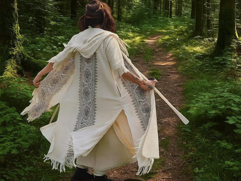 May include: Cream-colored hooded robe with intricate black geometric patterns along the front and sleeves. The robe has a fringed hem and is worn outdoors on a forest path. The person is holding a wooden staff.