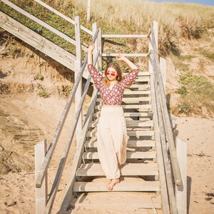 May include: A woman wearing a patterned purple and white crop top and cream-colored pants stands on a wooden staircase. She is wearing pink heart-shaped sunglasses and has her arms raised.