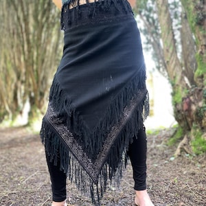 May include: A black fringed skirt with an embroidered border. The skirt is worn over black leggings. The image is taken outdoors with a blurred background of trees.