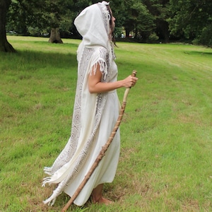 May include: A cream-colored hooded cloak with a geometric pattern in black and white, worn over a matching dress. The person is holding a wooden staff and standing in a grassy field.