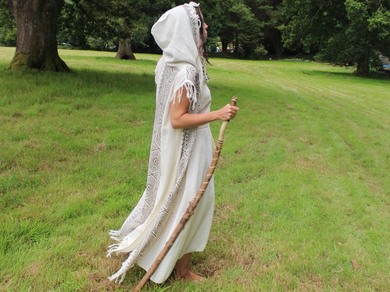 May include: A cream-colored hooded cloak with a geometric pattern in black and white, worn over a matching dress. The person is holding a wooden staff and standing in a grassy field.