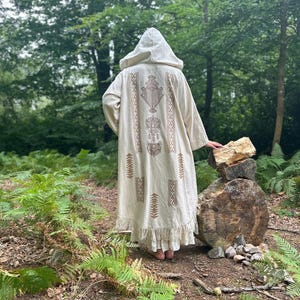 May include: A white hooded robe with brown embroidery featuring geometric patterns. The robe is worn by a person standing in a woodland setting.