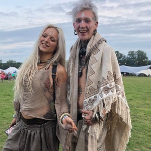 Op de afbeelding: Twee mensen die bohemian kleding dragen met witte gezichtsverf. De persoon links draagt een bruin gehaakt topje en broek. De persoon rechts draagt een witte sjaal met bruine geometrische patronen en franjes.