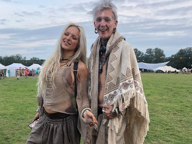 Op de afbeelding: Twee mensen die bohemian kleding dragen met witte gezichtsverf. De persoon links draagt een bruin gehaakt topje en broek. De persoon rechts draagt een witte sjaal met bruine geometrische patronen en franjes.