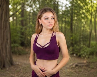 May include: A deep purple sports bra with a unique strappy design. The bra features a scoop neckline and a fitted silhouette. The model is standing in a forest setting with a blurred background of green trees and foliage.