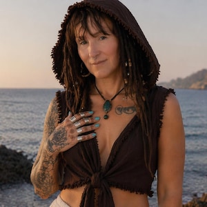 May include: A brown hooded top with a tie front. The sleeveless top has a fringed edge. The person is wearing multiple silver rings and a necklace with a turquoise pendant. The background is a seascape.