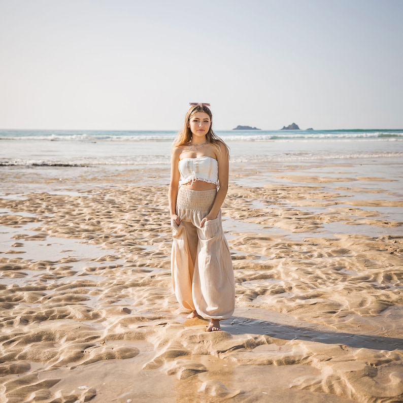 May include: A woman wearing a white crop top and beige wide-leg pants stands on a sandy beach. The ocean is in the background.