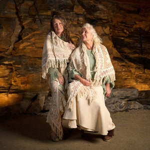 Op de afbeelding: Twee vrouwen die witte shawls met franjes dragen, staand voor een rotsachtige grot. De vrouw links heeft lang, donker haar en draagt een groen shirt. De vrouw rechts heeft wit haar en draagt een lichtgroen shirt.