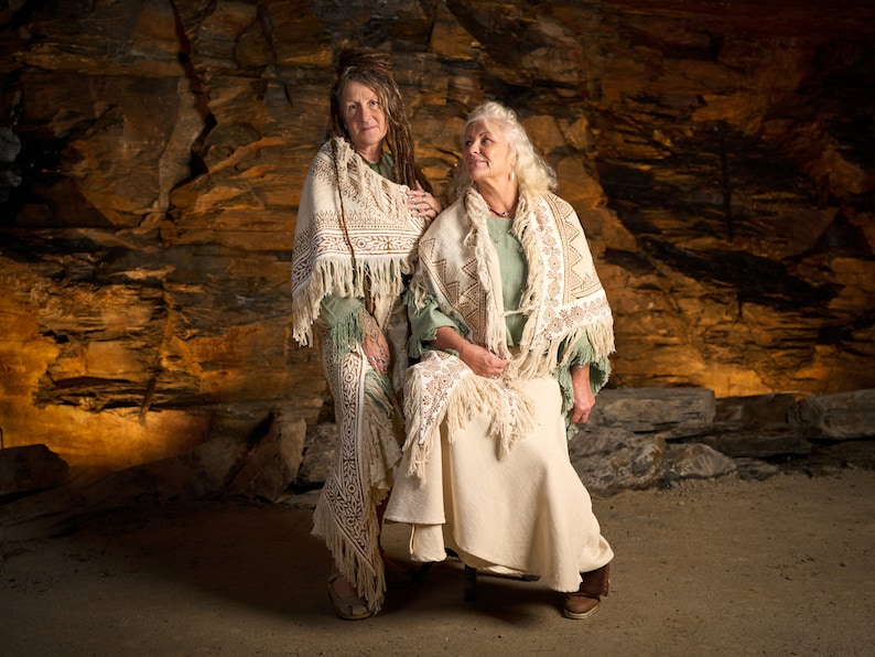Op de afbeelding: Twee vrouwen die witte shawls met franjes dragen, staand voor een rotsachtige grot. De vrouw links heeft lang, donker haar en draagt een groen shirt. De vrouw rechts heeft wit haar en draagt een lichtgroen shirt.