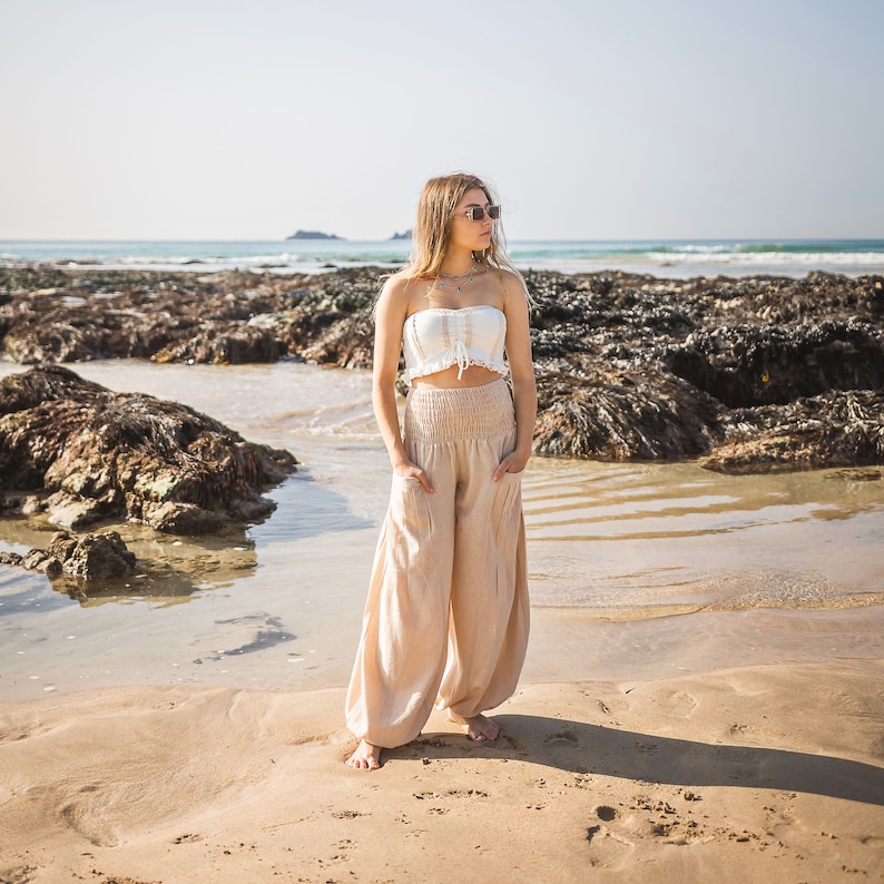 May include: A woman wearing a white crop top and beige wide-leg pants stands on a sandy beach with rocks and seaweed in the background.