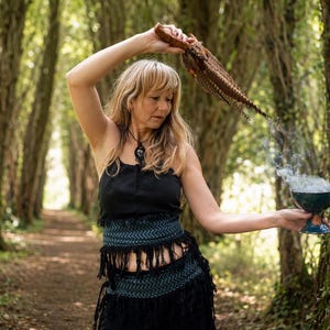 May include: A woman in a black outfit with a fringed skirt and a patterned belt holds a feathered object above her head. She holds a smoking teal bowl with a blue base. The background is a path through a forest.