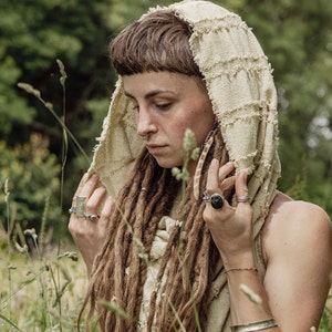 May include: A beige hooded garment with a textured, fringed design. The hood is worn over long, brown dreadlocks. The person is wearing multiple rings and bracelets. The background is a blurred view of green foliage.