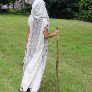 May include: A white hooded robe with a geometric pattern in dark gray, worn outdoors. The robe has fringe at the hem and sleeves. A wooden walking stick is held in the right hand. The person is standing in a grassy field.