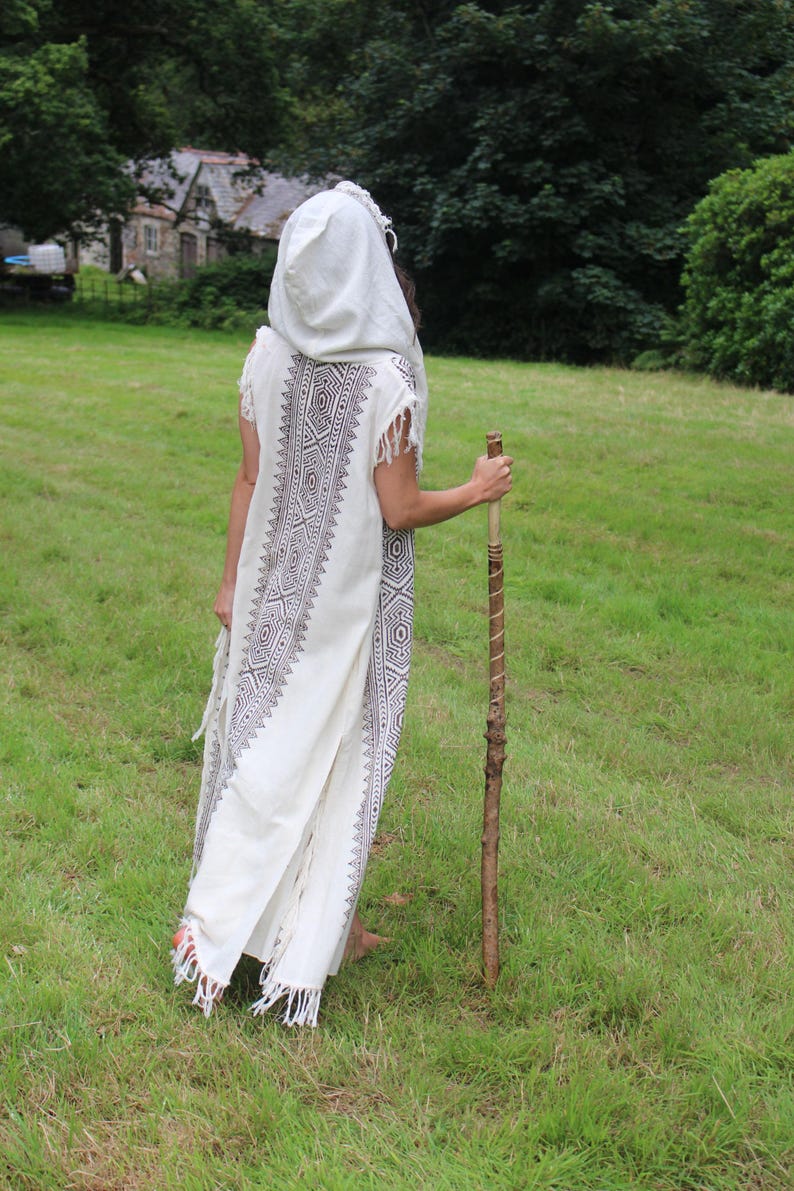 May include: A white hooded robe with a geometric pattern in dark gray, worn outdoors. The robe has fringe at the hem and sleeves. A wooden walking stick is held in the right hand. The person is standing in a grassy field.