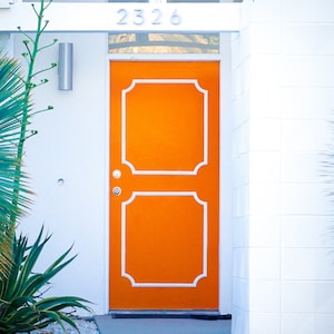 Peut inclure: Une porte d'entrée orange avec des bordures blanches et un design décoratif. La porte a un bouton de porte argenté et une poignée de porte argentée assortie. Le numéro de maison "2326" est au-dessus de la porte.