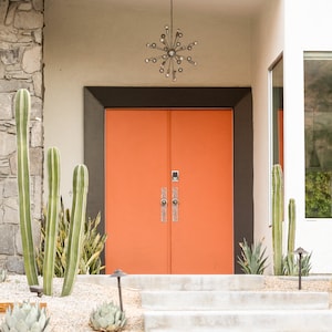 May include: A modern home with a bright orange double door entrance. The door has a dark brown frame and a silver door handle. The home has a white exterior and a stone wall with cactus plants.
