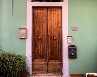 Könnte beinhalten: Eine zweiflügelige Holztür mit weißem Rahmen und einem dekorativen schmiedeeisernen Fenster darüber. Die Tür ist in eine grüne Wand mit einem Briefkasten und einer Hausnummertafel eingelassen.