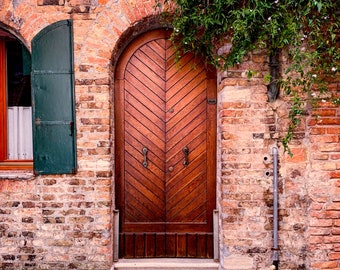 Könnte beinhalten: Eine Holztür mit Chevron-Muster, eingefasst in eine Backsteinmauer mit einem grünen Fensterladen links. Die Tür hat zwei Paneele und zwei Garnituren an Beschlägen. Die Tür wird von einem Backsteinbogen gerahmt.