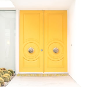 May include: A pair of bright yellow double doors with ornate brass door knobs. The doors are set in a white frame and have a yellow doormat.
