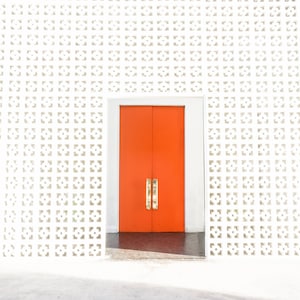 May include: A bright orange double door with gold handles is framed by a white doorway. The door is set against a white wall with a repeating geometric pattern. The floor is a dark grey colour.