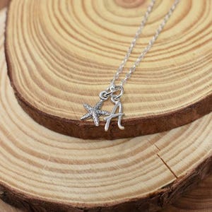 May include: Silver necklace with a starfish and letter A charm. The necklace is on a wooden surface.
