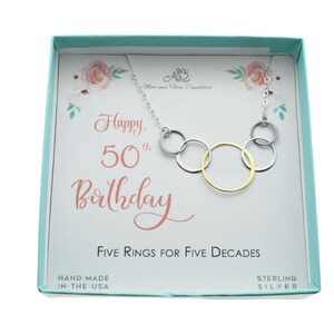 May include: A silver necklace with five interlocking rings, one gold ring, and a card that reads "Happy 50th Birthday" and "Five Rings for Five Decades". The card is in a light blue box with a floral design.