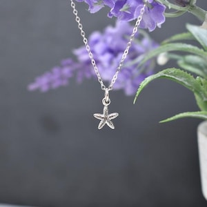 May include: A silver chain necklace with a small, detailed starfish pendant. The necklace is hanging against a purple and green background.