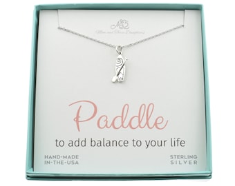 Könnte beinhalten: Eine silberne Halskette mit einem Paddel-Anhänger in einer hellblauen Geschenkbox. Die Box trägt den Text "Paddle to add balance to your life" und "Sterling Silver".