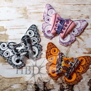 May include: Three embroidered felt butterfly shapes with lace details and a corset design in the centre. The butterflies are in black and white, pink and purple, and orange and brown.