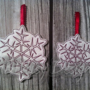 May include: Two white snowflake ornaments with burgundy stitching and a red ribbon for hanging. The ornaments are made of felt and have a detailed snowflake design. The background is a rustic wooden surface.