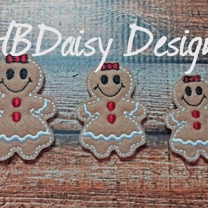 May include: Three felt gingerbread people ornaments with red bows, red buttons, and white icing. The ornaments are on a wood background with the text "HB Daisy Designs".