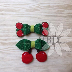 May include: Two felt cherry hair clips. The first clip is a bow with two red cherries and green leaves. The second clip is a pair of red cherries with green leaves.