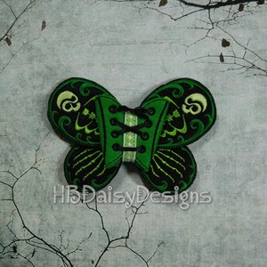 Op de afbeelding: Groen en zwart geborduurd vlinder-vormig patch met een schedel en gekruiste beenderen ontwerp. De patch heeft een korset-achtig ontwerp met zwarte en witte veters. De tekst "HBDaisyDesigns" is zichtbaar in de onderste helft van de afbeelding.