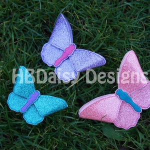 May include: Three glittery felt butterflies in pink, purple, and teal colours. The butterflies are arranged on a bed of green grass.