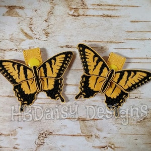 May include: Two yellow and black butterfly-shaped fabric clips with yellow buttons. The clips are on a white wood background.