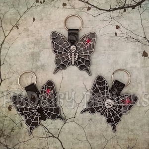 May include: Three black and white butterfly-shaped keyrings with a spiderweb design and a skull and crossbones motif. Each keyring has a red accent.
