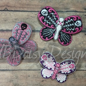 May include: Three embroidered butterfly patches featuring a pink, black and white colour scheme. The butterflies have a skeleton design with a skull on each wing. The patches are displayed on a wooden background.