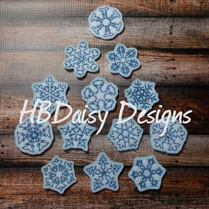 May include: Ten blue snowflake-shaped felt ornaments with white outlines, arranged in a pyramid shape on a brown wooden background. The text "HBD Daisy Designs" is visible in the center of the image.