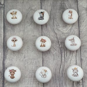 May include: Nine white round knobs featuring various animal illustrations. The animals include a lion, zebra, giraffe, meerkat, tiger, elephant, hippopotamus, and two different versions of a monkey. These knobs are ideal for adding a touch of whimsy to any room.