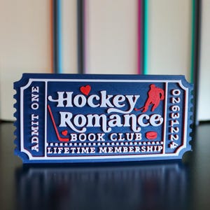 May include: A blue ticket-shaped item with white text reading "Hockey Romance Book Club". It features a red hockey player silhouette, a hockey stick, and a puck. The text "Admit One" is on the left side. The ticket is against a blurred background.