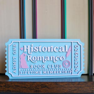 May include: A light blue ticket-shaped sign with the words "Historical Romance Book Club Lifetime Membership" in white. The sign has a pink silhouette of a couple and a feather illustration. The number "02631224" is printed on the right side.