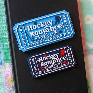 May include: Two ticket-shaped magnets with the text "Hockey Romance Book Club Lifetime Membership" on a black book. One magnet is light blue with a hockey player silhouette, and the other is navy blue with a red hockey player silhouette.