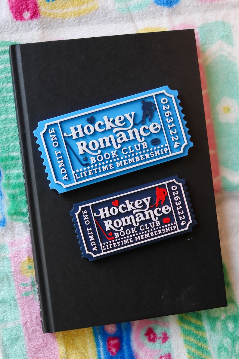 May include: Two ticket-shaped magnets with the text "Hockey Romance Book Club Lifetime Membership" on a black book. One magnet is light blue with a hockey player silhouette, and the other is navy blue with a red hockey player silhouette.
