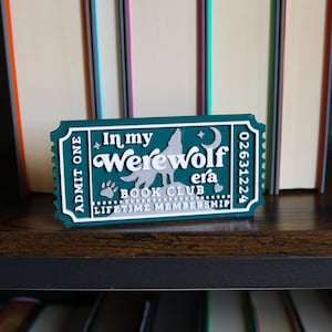 May include: A teal and white ticket-shaped sign that reads "In my Werewolf Era Book Club Lifetime Membership." The sign features a howling wolf, paw print, and moon design. The sign is on a dark wooden shelf with books in the background.