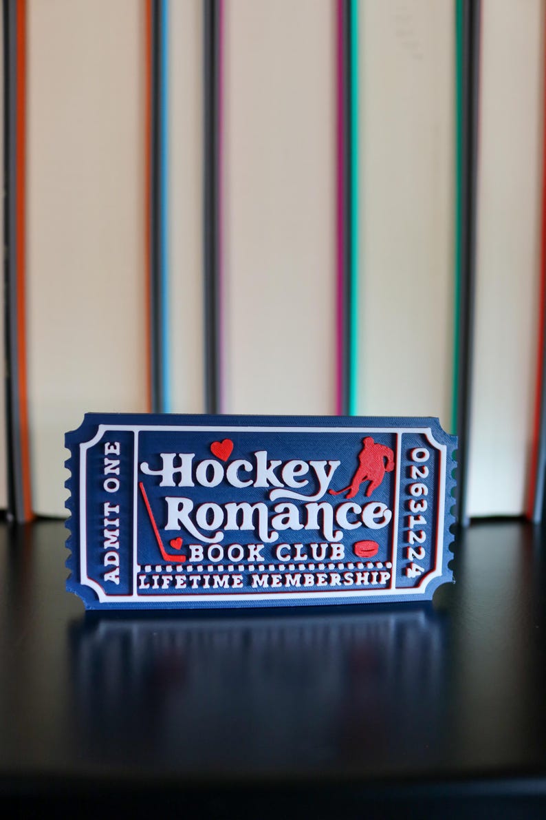 May include: A blue ticket-shaped item with white text reading "Hockey Romance Book Club". It features a red hockey player silhouette, a hockey stick, and a puck. The text "Admit One" is on the left side. The ticket is against a blurred background.